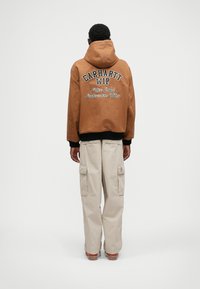 Καφέ κουκούλα φούτερ με μαύ�ρες μανσέτες και στρίφωμα, που διαθέτει τις λευκές κεντημένες επιγραφές "CARHARTT WIP" και "Jacket Anniversary Edition". Μπεζ cargo παντελόνι.