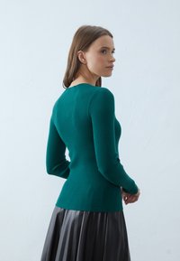 Teal ribbestrikket genser med lange ermer og rund hals, kombinert med et mørkt, plissert skjørt. Materialet ser mykt ut med en tettsittende design.