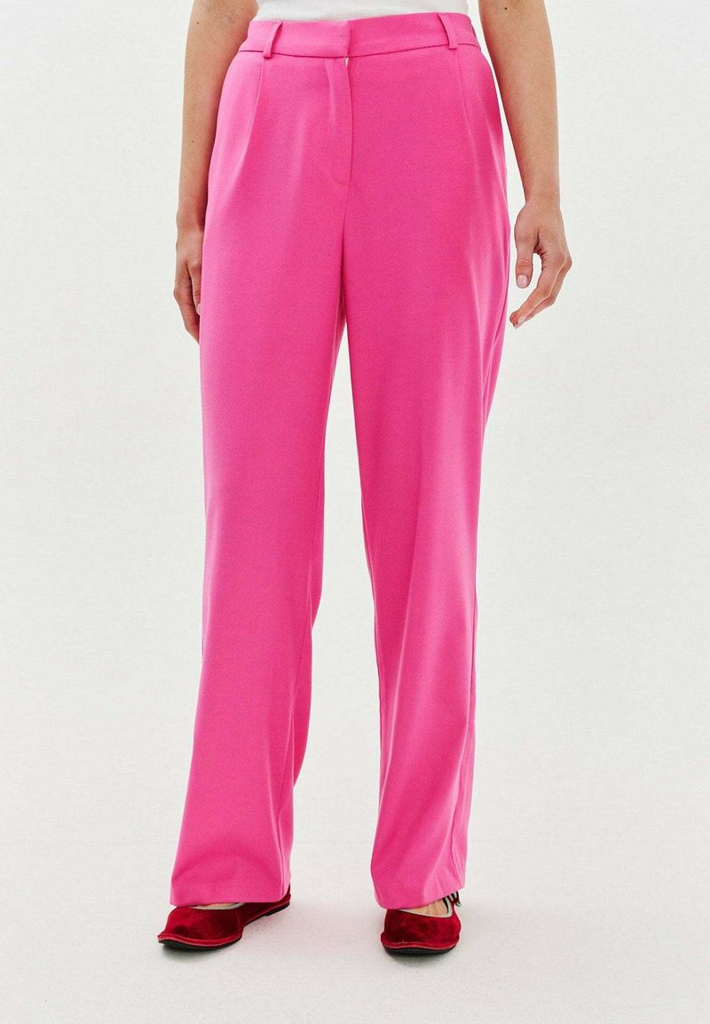 Różowe spodnie od garnituru o gładkiej fakturze, prostym kroju i subtelnymi plisami. W połączeniu z czerwonymi płaskimi butami.