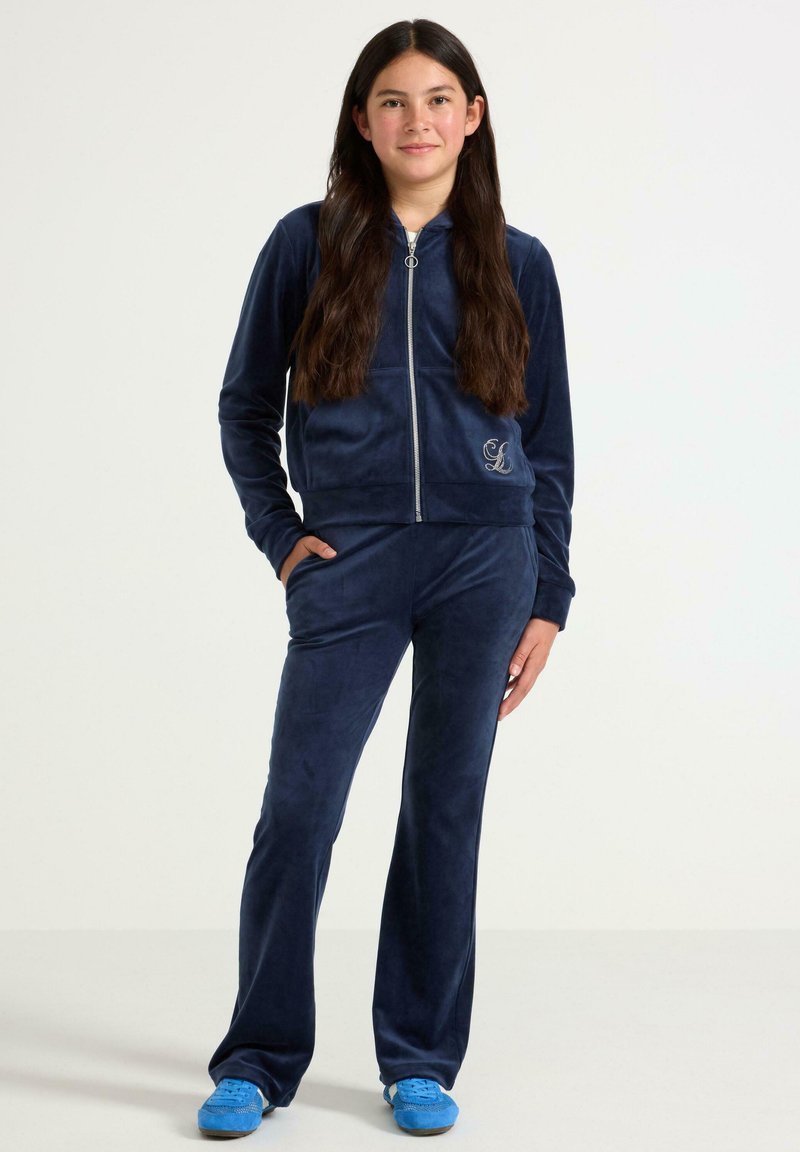 Navyblauer Velours-Trainingsanzug mit einer Jacke mit Reißverschluss, Taschen und besticktem Logo, kombiniert mit passender Hose und blauen Sneakers.