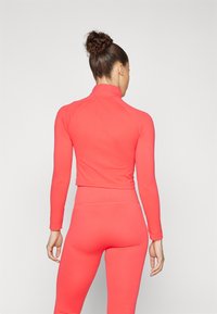 Korallfarget langermet treningsgenser med høy krage, tettsittende design og glatt tekstur. Kombinert med matchende leggings, som fremhever et sammenhengende utseende.