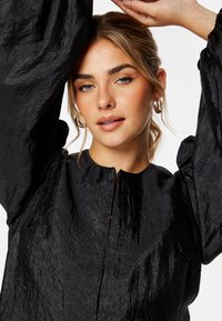 Zwarte, gestructureerde blouse met een rits aan de voorkant, hoge halslijn en volumineuze mouwen. Heeft een gekreukt stofpatroon en een subtiele glans.