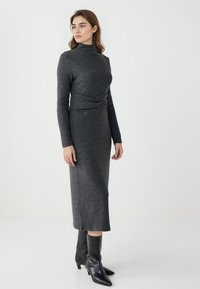 Graues langärmliges Jersey-Kleid mit hohem Ausschnitt und geknotetem Taillendetail, kombiniert mit schwarzen spitzen Ankle Boots. Glatte Textur.