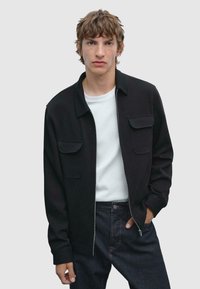 Veste noire zippée avec deux poches poitrine, associée à un t-shirt blanc et un jean foncé. La veste a une texture lisse et un design moderne.