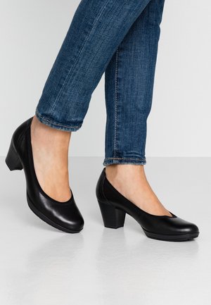 Jambes en jean denim bleu portant des chaussures en cuir noir à talons moyens et bouts arrondis, debout sur une surface blanche brillante.