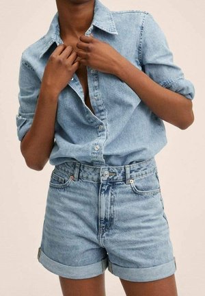 Person iført lys blå denimskjorte med opkrullede ærmer og matchende højttaljede denimshorts, som justerer skjortens knapper.