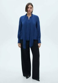 Blusa blu a maniche lunghe con patta frontale e cravatta attaccata, abbinata a pantaloni neri a gamba larga e décolleté neri a punta.