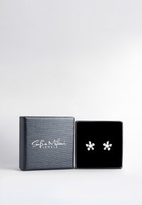 Silverfärgade blommörade örhängen på en svart sammetskudde, placerade i en texturerad svart smyckeskrin med vit logotext.