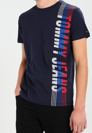 Navy blue short-sleeve shirt with vertical red, white, and blue "TOMMY JEANS" text on the front, worn by a person with hands in pockets.