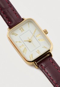 Stačiakampio formos aukso spalvos laikrodis su balta ciferblatu, aukso romano skaitmenimis ir bordo spalvos odiniu dirželiu su raudonomis siūlėmis.