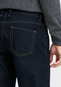 Mørkeblå denimjeans med en enkelt baklomme, kontrastsøm og slim fit, vist mot en grå genser.