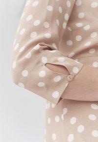 Beige Bluse mit großen weißen Punkten; verfügt über einen knöpfbaren Ärmel mit Schlitz. Der Stoff wirkt weich mit einer glatten Textur.
