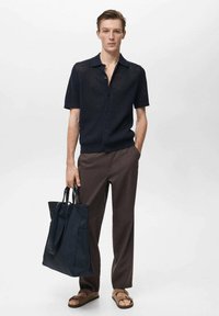 Camicetta in maglia blu navy a maniche corte con chiusura a bottoni, abbinata a pantaloni marroni. Il modello tiene in mano una borsa tote scura e indossa sandali marroni.