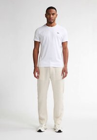 White short-sleeve t-shirt with a crew neck, paired with beige, loose-fitting pants. Worn with white sneakers, featuring a subtle logo on the chest.
