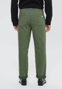 Pantalon en coton vert avec une coupe droite, comportant deux poches arrière et des poignets retroussés, associé à des chaussures noires. Design simple et fonctionnel.