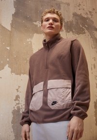 Hnedý fleece pulóver s polovičným zipsom, ktorý má dve veľké ružové vrecká a čierne logo Nike na pravom vrecku. Textúrovaná látka.