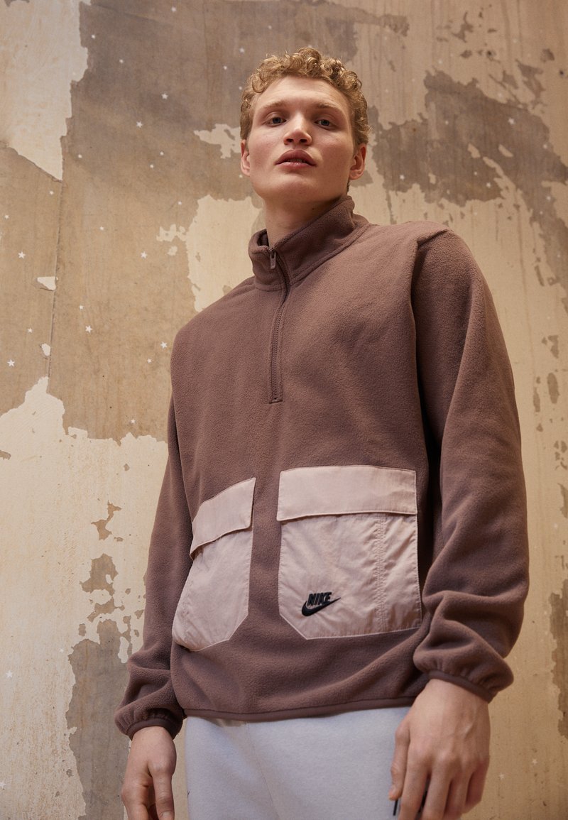 Hnedý fleece pulóver s polovičným zipsom, ktorý má dve veľké ružové vrecká a čierne logo Nike na pravom vrecku. Textúrovaná látka.