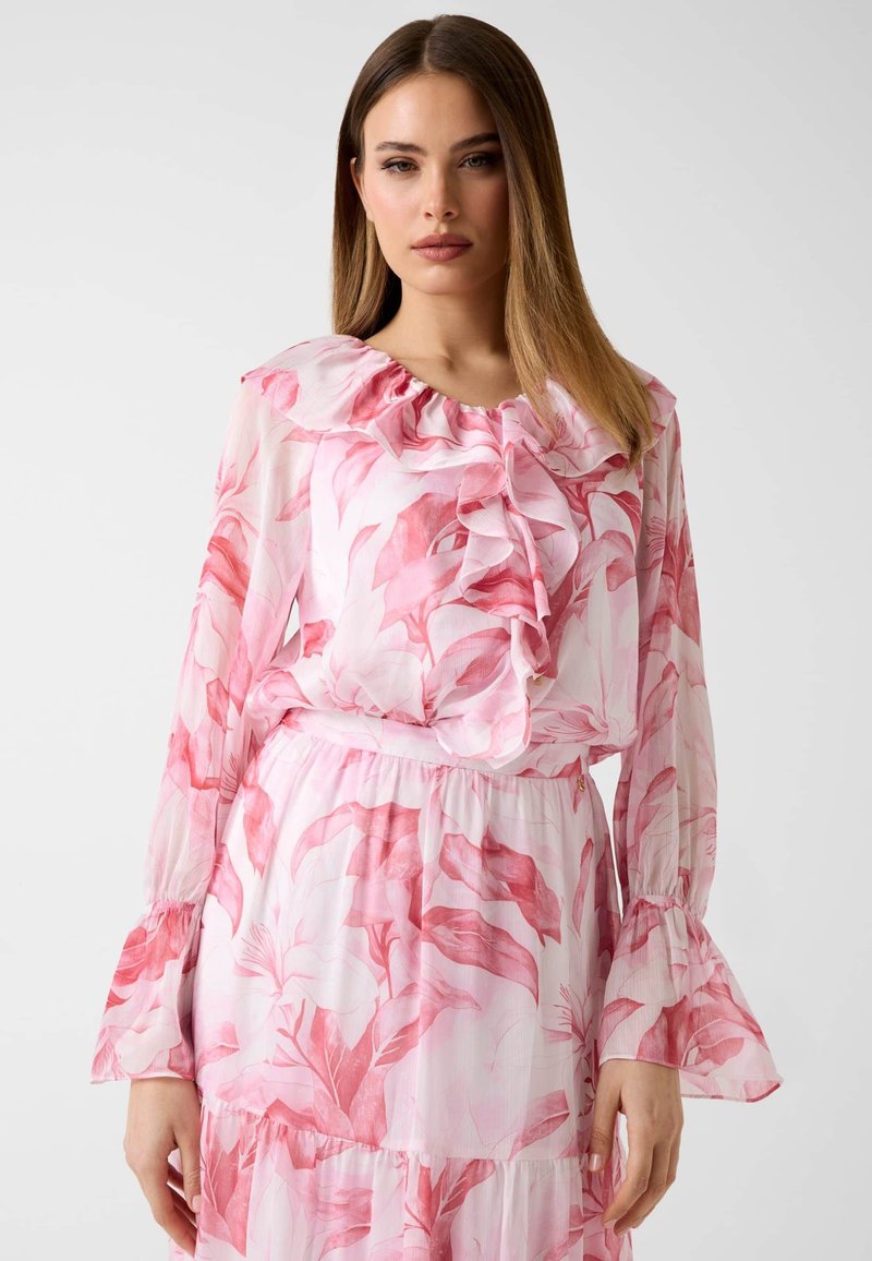 Femme aux cheveux raides portant une robe blanche transparente avec un imprimé floral rose, un col à volants et des manches évasées sur fond uni.