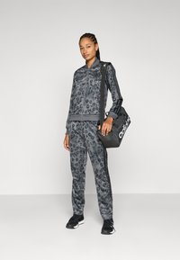 Grå träningsoverall med leopardsmönster, bestående av en zip-up jacka och matchande byxor. Modellen håller en svart adidas väska med ränder och logotypaccent.