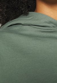 Capelli scuri e ricci e collo visibili sopra il colletto di un indumento in tessuto verde, con dettagli di trama e cuciture.