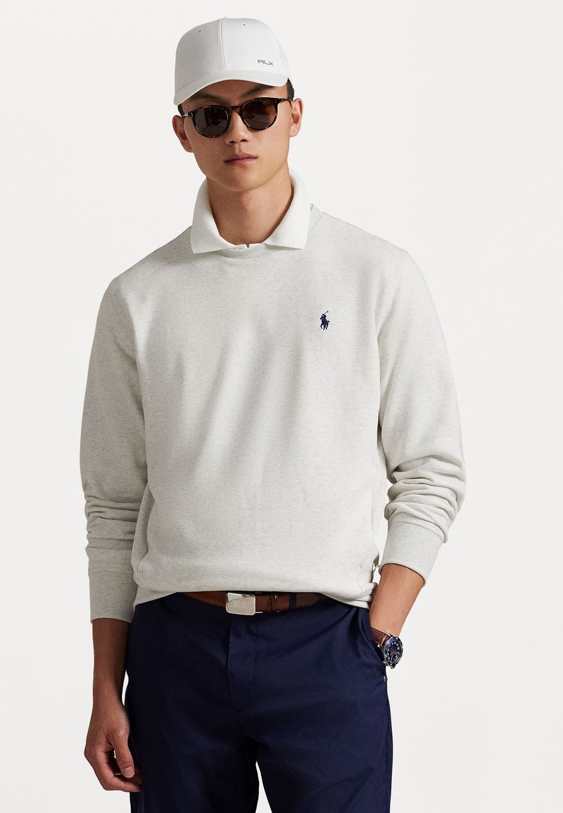 Lichtgrijze pullover met een witte kraag en een marineblauw logo. Gecombineerd met marineblauwe broek en een bruine riem. Accessoires zijn een witte pet en een zonnebril.
