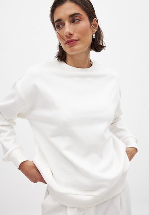 Sudadera blanca hecha de un tejido suave, con cuello redondo y puños acanalados. Corte holgado con bolsillos laterales. Textura suave, diseño minimalista.