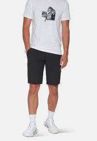Svarta shorts tillverkade av stretchmaterial, med sidofickor och en logotyp-patch. Kombinerat med en vit grafisk T-shirt och ljusa sneakers.