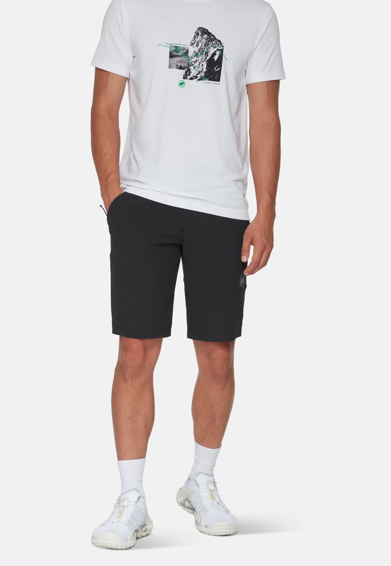 Svarta shorts tillverkade av stretchmaterial, med sidofickor och en logotyp-patch. Kombinerat med en vit grafisk T-shirt och ljusa sneakers.