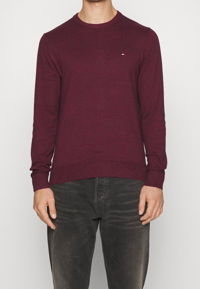 Weinroter Pullover mit Rundhalsausschnitt, Strickstoff, lange Ärmel, gerippte Bündchen und Saum, dezentes Logo auf der Brust, kombiniert mit dunkler Jeans.