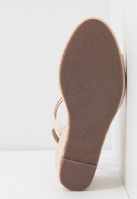 Semelle de sandale compensée beige présentée verticalement contre des murs blancs, montrant une semelle en caoutchouc texturée et le marquage taille 37.