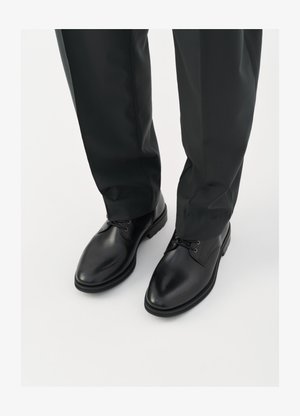Fekete bőr cipők sima felülettel, kerek orral és fűzős kivitelben, sötét nadrággal párosítva, fehér háttér előtt.