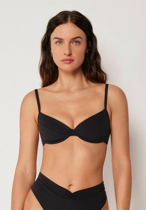Mujer con cabello largo y oscuro que lleva un top de bikini negro con aro y braguitas de bikini negras de talle alto sobre un fondo liso.