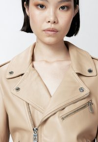 Cazadora de moto de cuero marrón claro con cuello con muescas, cremallera plateada, botones a presión y costuras decorativas. Incluye un bolsillo en el pecho con cremallera.