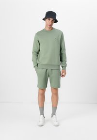 Grünem Baumwoll-Sweatshirt mit gerippten Bündchen, kombiniert mit passenden Shorts. Weiße Sneakers und graue Socken vervollständigen den Look. Ein schwarzer Bucket Hat betont den Stil.