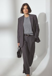 Pelēks piegriezums uzvalks ar platām biksēm un garu žaketi, saskaņots ar pelēku blūzi. Aksesuāri ietver melnu rokassomu.