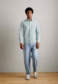 Cămașă cu mânecă lungă în dungi verzi deschis și albe, blugi din denim albastru și adidași cu accente verzi, pe un fundal neutru.