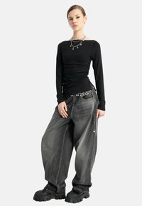 Młoda kobieta w czarnej bluzce z długim rękawem, oversize'owych, wyblakłych szarych dżinsach, masywnych czarnych butach oraz srebrnej biżuterii i pasku z motywem gwiazdy.