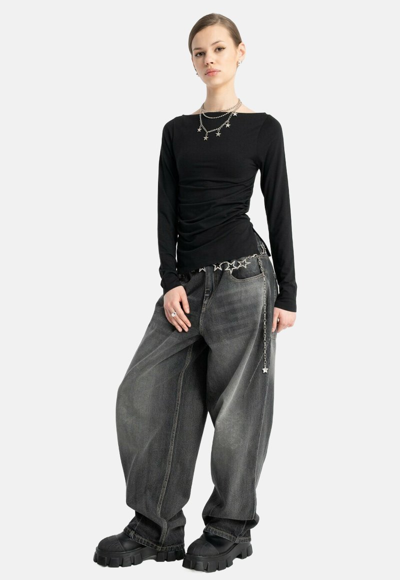 Młoda kobieta w czarnej bluzce z długim rękawem, oversize'owych, wyblakłych szarych dżinsach, masywnych czarnych butach oraz srebrnej biżuterii i pasku z motywem gwiazdy.