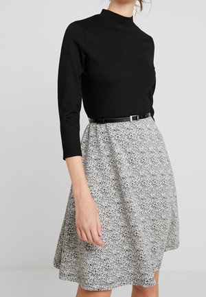 Haut noir à manches longues associé à une jupe évasée avec un motif texturé noir et blanc, cintrée à la taille par une ceinture noire.