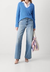 Woman wearing a blue knit sweater over a white shirt, light blue wide-leg jeans, nude heels, and holding a cream bag with a pink and blue scarf.