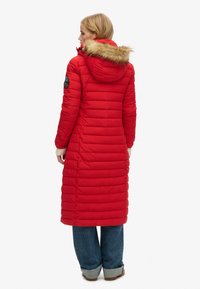 Rød polstret lang frakke med aftagelig faux fur hætte, ribbede manchetter og quiltet tekstur. Har en sort patchdetalje på ærmet.