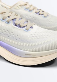 Gros plan sur des chaussures de course beige et lavande avec une tige en mesh texturé, des lacets beiges, une semelle intermédiaire beige rembourrée intitulée "HyperTech" et une semelle extérieure noire.