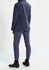 Person wearing a navy blue jacket and matching pants, standing with back facing the viewer, wearing black athletic shoes with pink accents.