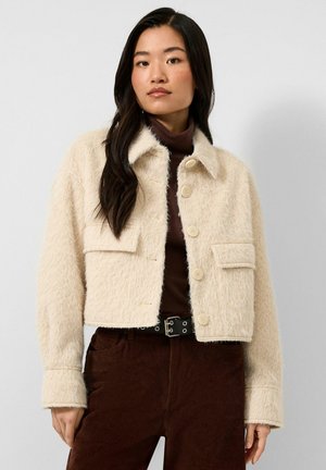 Jeune femme aux longs cheveux foncés porte une veste crème duveteuse boutonnée sur un col roulé marron et un pantalon marron foncé avec une ceinture noire.