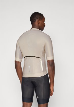 Maillot de cyclisme beige clair à manches courtes, en tissu texturé, avec une poche zippée à l'arrière et des détails réfléchissants. Associé à un cuissard de cyclisme noir rembourré.