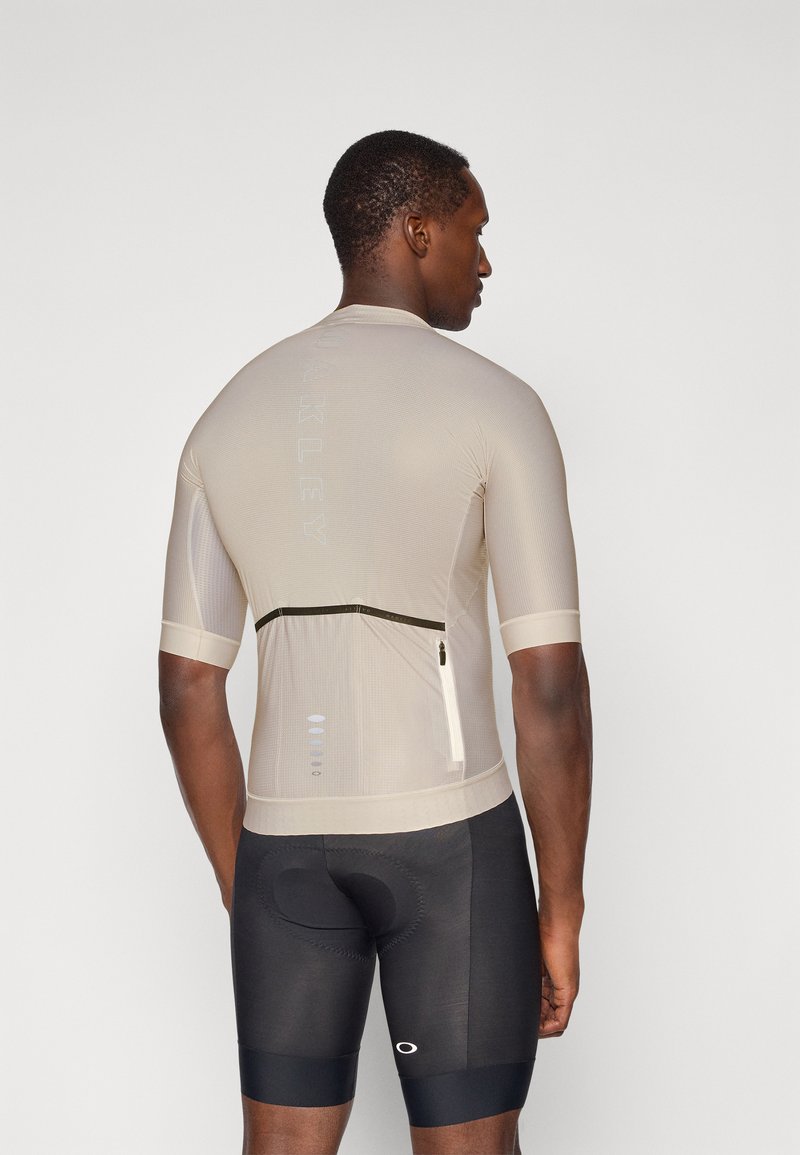 Helle beige Fahrradtrikot mit kurzen Ärmeln, strukturiertem Stoff, rückseitiger Reißverschlusstasche und reflektierenden Details. Kombiniert mit schwarzen gepolsterten Fahrradshorts.