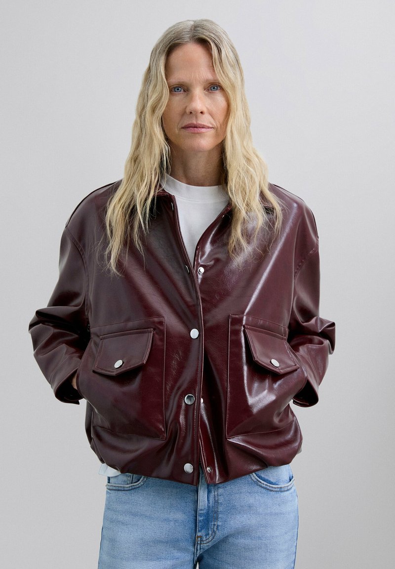 Veste en simili cuir bordeaux avec une coupe décontractée, dotée de boutons-pression argentés, de deux poches avant et d'une texture légèrement brillante.