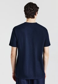 Uomo con capelli ricci scuri che indossa una camicia a maniche corte blu navy semplice, mostrato di spalle su uno sfondo chiaro e semplice.