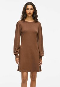 Robe longue marron à manches longues avec une coupe décontractée, texture douce, encolure ronde et détails froncés au niveau des épaules. L'ourlet tombe au-dessus du genou.