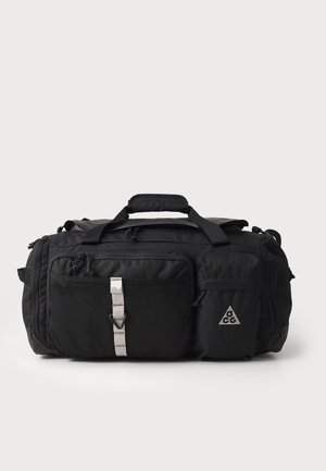 Sac de sport noir avec une poche en mesh texturé, des accessoires en métal argenté et des bretelles réglables. Présente un logo en triangle distinctif et un tissu durable.